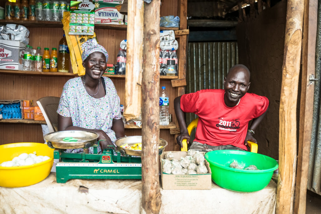 A woman and a man sitting inside their shop selling vegetables and other household supplies. 